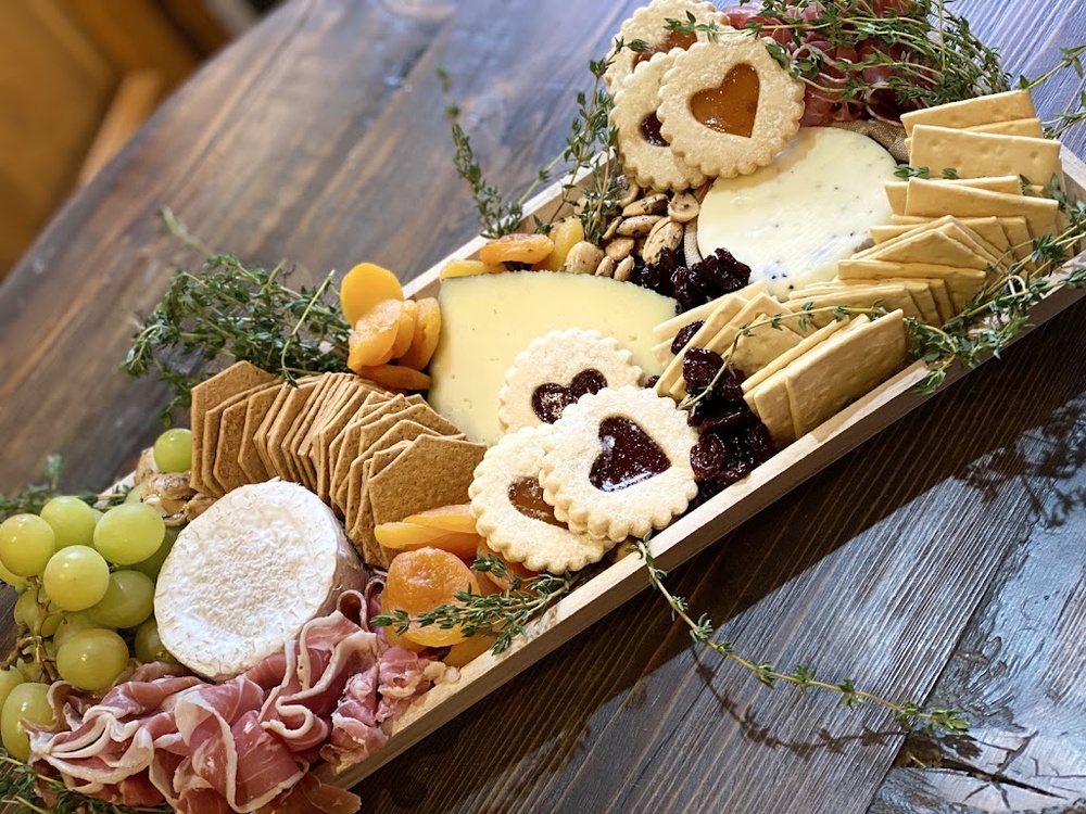 A charcuterie board in a wooden trough: prosciutto, brie, aged cheddar, Linzer cookies, dried apricots, grapes, and fresh thyme