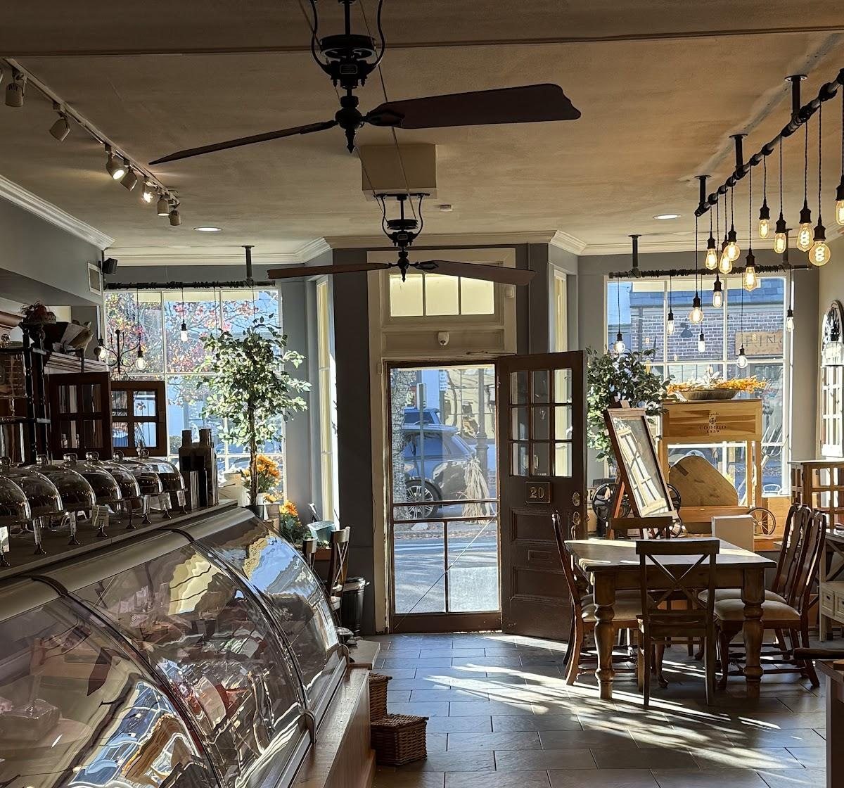 Inside the shoppe: the curved glass display case, farmhouse tables, Edison bulbs, and the front door marked 20 in afternoon sun